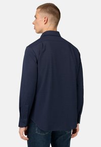 Modra srajca z dolgimi rokavi in gumbastim ovratnikom, ki ima teksturirano blago in zaobljen rob. Rokavi so zaključeni z gumba.