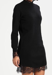 Svart stickad klänning med långa ärmar, med hög hals och spetsdetaljer vid ärmarna och fåll, vilket skapar en lagertextur.