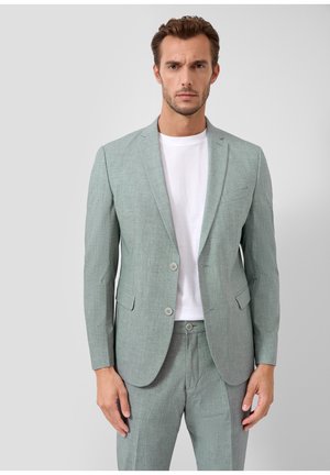 Man draagt een lichtgroed pak met een witte T-shirt met ronde hals en staat tegen een effen lichtgrijze achtergrond.