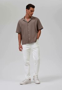 Hombre con una camisa de botones marrón de manga corta, pantalones blancos y zapatillas blancas, de pie con una mano en el bolsillo, mirando hacia un lado.