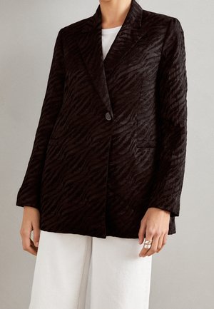 Vrouw draagt een gestructureerde zwarte single-button blazer met zebra-achtig patroon over een witte top en witte broek, staand tegen een grijze achtergrond.