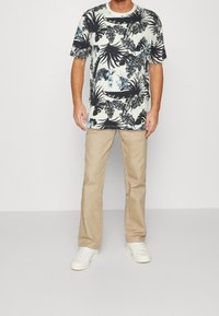 Košile s krátkým rukávem, světle modrá s bílým tropickým potiskem a tmavě zelenými listy; spárována s béžovými kalhotami a bílými teniskami.