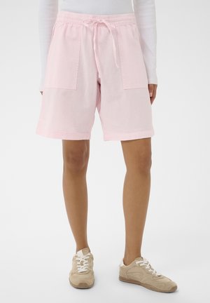 Person iført lyserøde shorts med snor og store forlommer samt beige sneakers, stående foran en ensfarvet baggrund.