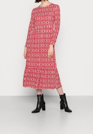 Kobieta w czerwono-białej sukience midi z długim rękawem i geometrycznym wzorem, w czarnych botkach na obcasie, stojąca na tle jednolitego tła.
