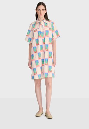 Frau trägt ein knielanges, kurzärmliges Kleid mit buntem kariertem Muster und beigen Slippern, steht vor einem weißen Hintergrund.