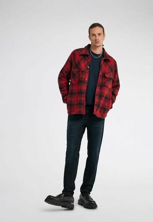 Giovane uomo con giacca a quadri rossa e nera, jeans scuri, scarpe nere spesse, in piedi con le mani in tasca su sfondo bianco.