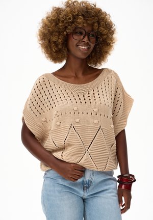 Kobieta o kręconych włosach, nosząca duże okulary, beżowy sweter wytarty z wzorami, jasnoniebieskie dżinsy i czerwone bransoletki, uśmiechająca się z ręką w kieszeni.