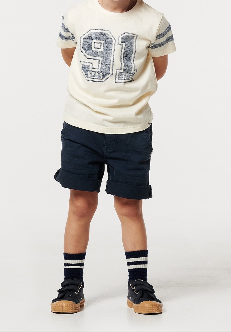 Enfant portant un t-shirt crème avec le numéro 91, un short bleu marine, des chaussettes rayées et des baskets noires, debout les mains derrière le dos.