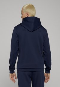 Tmavě modrá mikina s kapucí s texturovanými bočními panely a vyraženým logem na zádech. Hladká látka s žebrovanými manžetami a lemem.