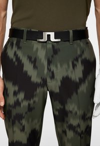 Svart läderbälte med silverfärgad hårdvara, i kombination med byxor i grönt och svart med skarpa, abstrakta mönster.