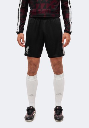 Persona con pantalones cortos negros de fútbol del Liverpool FC, calcetines blancos hasta la rodilla con el logo de Adidas, botas negras Adidas y una camiseta de manga larga con estampado.