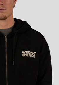 Černá mikina na zip vyrobená z měkké tkaniny, s přední kapsou a krémovým vyšitým logem "WRONG FRIENDS" na levém prsu.