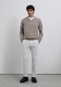 Beige V-hals trui, wit T-shirt eronder, lichtgekleurde broek en zwarte schoenen. Model staat rechtop, handen in de zakken, tegen een effen achtergrond.