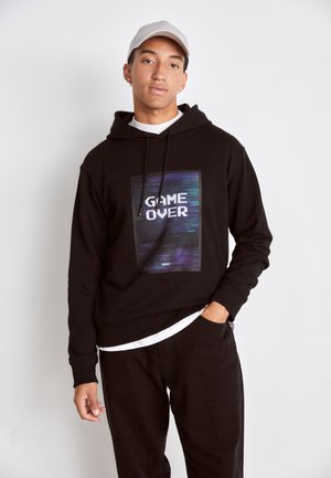 Felpa nera con cappuccio caratterizzata da una grafica rettangolare che mostra "GAME OVER" in un font bianco pixelato, sovrapposta a uno sfondo scuro e colorato.
