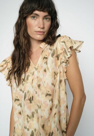 Femeie cu păr lung, ondulat și șaten, purtând un top bej cu mâneci scurte și volănașe, cu modele abstracte roz și maro, privind spre cameră.