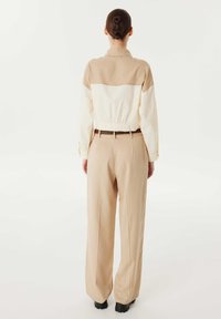 Giacca beige e bianca corta con vita elastica, maniche lunghe e polsini a bottone, abbinata a pantaloni beige larghi e scarpe nere.
