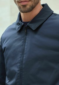Jachetă bleumarin cu fermoar în față și guler, realizată dintr-un material neted, având o strălucire subtilă și detalii de cusătură curate.