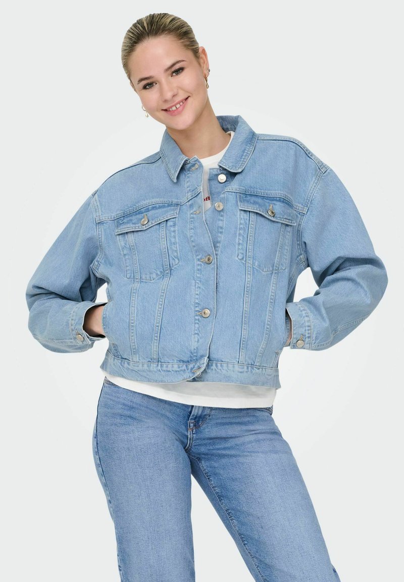 Lächelnde Frau trägt hellblaue Jeansjacke und Jeans, steht mit den Händen in den Jackentaschen vor einem einfarbigen Hintergrund.