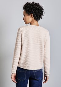 Světle růžový úpletový svetr s kulatým výstřihem a dlouhými rukávy, kombinovaný s tmavě modrými džíny. Viditelné švy na džínech.