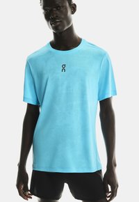 Tânăr stând în picioare, purtând un tricou sportiv bleu deschis și pantaloni scurți negri, pe un fundal alb.