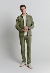 Olijfgroene jack met twee borstzakken, gecombineerd met bijpassende broek en een beige shirt, afgewerkt met lichtgekleurde sneakers.