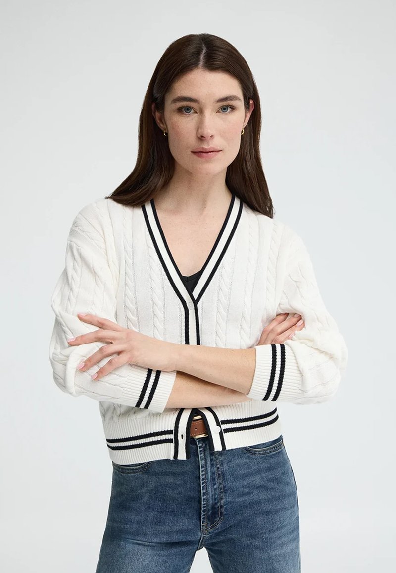 Frau mit langen braunen Haaren, die eine weiße, kabelgestrickte Strickjacke mit schwarzen Streifen und blaue Jeans trägt, die Arme verschränkt, vor einem schlichten Hintergrund.