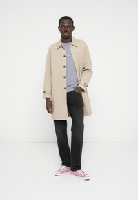 Μπεζ παλτό τύπου trench με καφέ κουμπιά, γκρι μπλουζάκι, μαύρο παντελόνι και ροζ αθλητικά παπούτσια. Το παλτό έχει ίσια γραμμή και μακριά μανίκια.