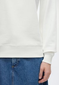 Λευκό φούτερ με μακριά μανίκια, ριμπ μανσέτες και στρίφωμα, φορεμένο με μπλε τζιν, που δείχνει ένα άνετο, καθημερινό ντύσιμο.