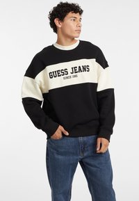 Sweatshirt in Schwarz und Creme mit Color-Blocking, versehen mit dem Schriftzug "GUESS JEANS SINCE 1981". Hergestellt aus weichem Stoff mit gerippten Bündchen und einem Rundhalsausschnitt.