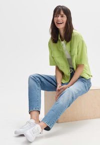 Maglietta verde lime a maniche corte con due tasche frontali, abbinata a jeans blu chiaro e sneakers bianche, seduta su un blocco di legno chiaro.