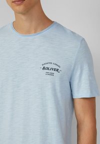 Jasnoniebieska, krótko rękawowa koszulka z miękkiego, lekkiego materiału. Posiada okrągły dekolt i czarny nadrukowany logo na piersi.