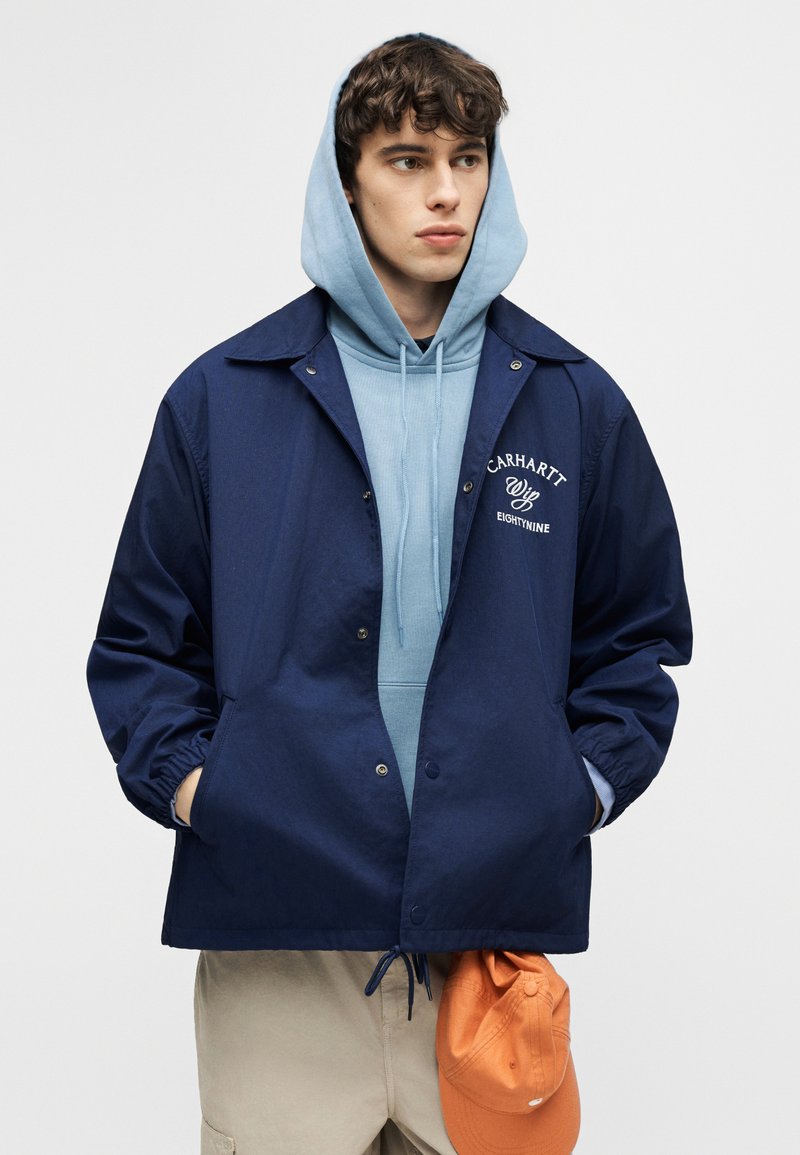 Giovane uomo con una felpa con cappuccio azzurro chiaro sotto una giacca Carhartt blu navy, che tiene in mano un cappellino arancione e guarda di lato.
