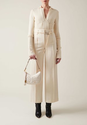 Maglione crema con colletto, pantaloni crema a vita alta con cintura, tiene una borsa a tracolla bianca trapuntata con dettagli dorati, abbinata a stivaletti neri.