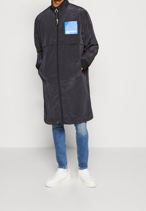 Homme portant un long manteau noir zippé avec un logo carré bleu, un jean bleu et des baskets blanches, debout les mains dans les poches.