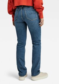 Blå denimbukser med en tætsiddende pasform, der har to baglommer og kontrastsyet, sammen med en rød kortærmet sweatshirt og lyse sneakers.