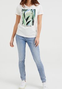 Bílá tričko s zeleným botanickým potiskem, obsahující text "La Dolce Vita"; v kombinaci se světle modrými džíny a bílými teniskami. Jednoduchý design.