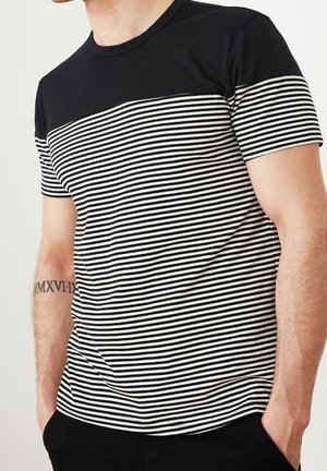 Man draagt een zwart-wit horizontaal gestreept t-shirt met zwart bovenstuk en mouwen, handen in de zakken, tatoeage op de linker onderarm.