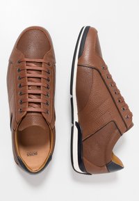 Baskets en cuir marron avec surfaces texturées, bout rond et accents noirs contrastants. Elles possèdent une semelle en caoutchouc blanche et un design à lacets.