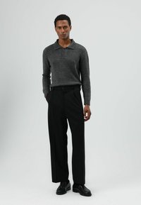 Donkergrijs gebreid poloshirt met kraag en knoopsluiting, gecombineerd met hoge zwarte broek en zwarte schoenen, staand tegen een lichte achtergrond.