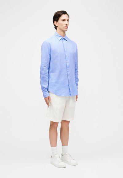 Άνδρας που στέκεται φορώντας ανοιχτό μπλε πουκάμισο με μακριά μανίκια κουμπωμένο, άσπρο σορτς μέχρι το γόνατο, άσπρες κάλτσες και άσπρα αθλητικά παπούτσια, μπροστά σε απλό φόντο.