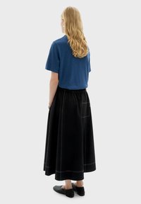 Chemise bleue à manches courtes, jupe plissée noire avec poches et coutures visibles, portée avec des chaussures plates noires. Cheveux longs, blonds et ondulés.