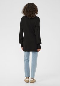 Pulover negru cu mâneci lungi, cu croială lejeră și manșete striate, asortat cu blugi bleu deschis și pantofi bej, purtat din spate.