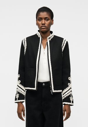 Femme portant une veste noire avec un galon brodé blanc et un chemisier blanc en dessous, debout devant un fond uni.