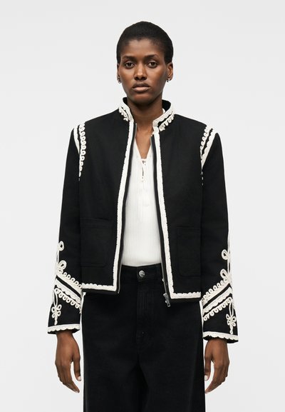 Femme portant une veste noire avec un galon brodé blanc et un chemisier blanc en dessous, debout devant un fond uni.