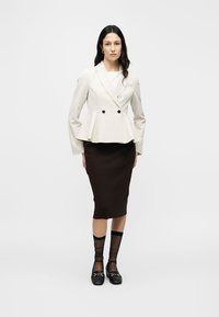 Blazer blanc à basque avec double boutonnage, superposé à un haut clair, associé à une jupe crayon marron côtelée et des ballerines noires avec accents.
