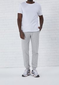 Bílý tričko s krátkým rukávem, šedé tepláky a stříbrné sportovní boty. Neformální outfit na pozadí světlé cihlové zdi.