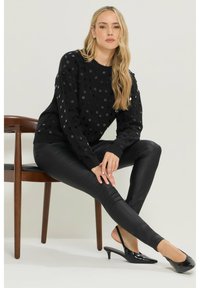 Czarny sweter z wypukłymi kropkami, zestawiony z błyszczącymi czarnymi legginsami. Modelka nosi czarne buty na obcasie typu slingback. Neutralne tło i drewniane krzesło.