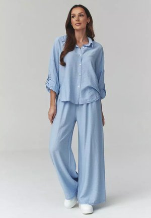 Kobieta ubrana w luźną, jasnoniebieską koszulę na guziki z podwiniętymi rękawami oraz pasujące szerokie spodnie, stojąca na jednolitym szarym tle.