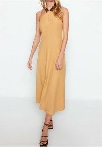 Kvinna klädd i en ärmlös klänning i senapsgul halterneck-modell med en nyckelhålsringning och bruna sandaler med hög klack och remmar.