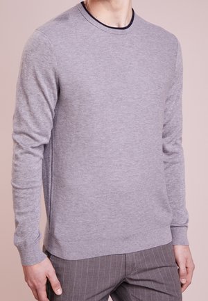 Muž nosící světle šedý pletený svetr s texturou a žebrovanými manžetami, doplněný šedými kalhotami s proužky, na jednobarevném pozadí.